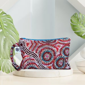 May include: A rectangular pouch with a colourful, dotted pattern in red, white, blue, and orange. The pouch has a blue zip and a matching handle. The design is reminiscent of Aboriginal art. The pouch is displayed on a white plinth.
