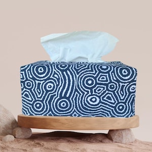 May include: A navy blue tissue box cover featuring a white, abstract design. The fabric box rests on a round, wooden base supported by small stones. White tissues are seen emerging from the top.