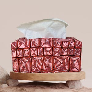 May include: A red tissue box cover featuring a white geometric design, placed on a wooden stand. White tissues protrude from the top. The cover is supported by two small stones on a sand-coloured surface.