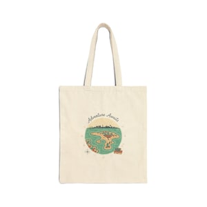 May include: A cream-coloured tote bag featuring a map design and the words "Adventure Awaits." The map shows an island with a city skyline, surrounded by green water. A small train is also depicted on the map.