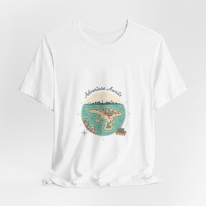 May include: White t-shirt with a circular island map design. The map includes a compass, a boat, and the words "Adventure Awaits". The island is in green, brown, and blue. The t-shirt is a casual wear item.