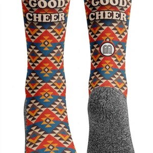 May include: A pair of crew socks featuring a geometric pattern in red, blue, yellow, and white. The text BE OF GOOD CHEER is printed in white on the upper portion of the socks. The heel and toe sections are a solid heather gray color.