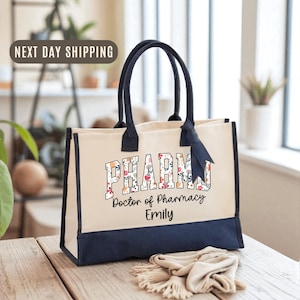 May include: Cream-colored tote bag with navy blue accents and handles. The bag features the word "PHARM" in a floral design, along with "Doctor of Pharmacy" and a name. A navy blue ribbon is tied to the handle.