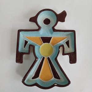 May include: A ceramic dish in the shape of a stylized bird figure. The dish features a light blue interior with brown, yellow, and dark brown geometric designs. The bird's head has a circular indentation, and the wings are outstretched.