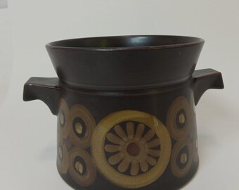 May include: A dark brown ceramic bowl with two side handles. The bowl features a decorative pattern of concentric circles and a central floral design in shades of brown and tan.