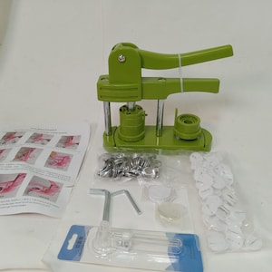 May include: A light green button making machine with a manual lever, accompanied by a set of metal button shells, white plastic backings, a clear plastic circle cutter tool, and an instructional sheet with diagrams for assembly and operation.