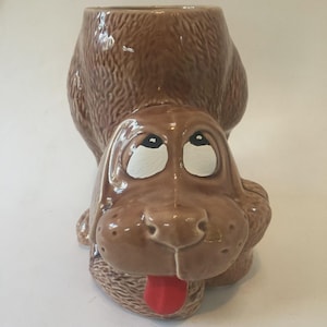 Pode incluir: Um vaso de cerâmica castanho em forma de cão com uma língua vermelha e olhos grandes e expressivos. O vaso tem uma superfície texturizada e uma abertura larga no topo. A cara do cão é pintada com detalhes brancos e pretos.