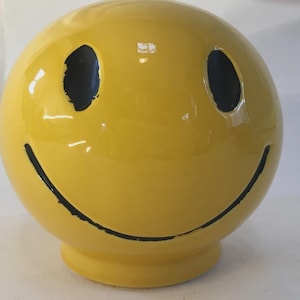 May include: A yellow ceramic smiley face sphere with black eyes and a curved black smile. The sphere has a glossy finish and a small, circular base. The design is simple and cheerful, evoking a sense of happiness.