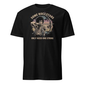 May include: Black t-shirt featuring a graphic of a musician with a bow and arrow. The image is surrounded by a circular design with the text "SOME ROCK STARS" above and "ONLY NEED ONE STRING" below.