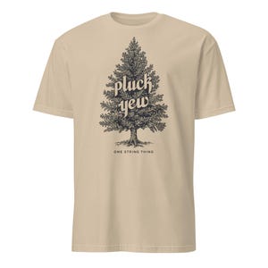 May include: A tan t-shirt featuring a detailed black illustration of a pine tree with the words "pluck yew" in a stylized font. Below the tree, the text "ONE STRING THING" is printed in a smaller font.