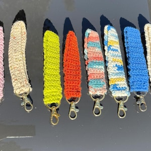 May include: A collection of eight handmade crochet wrist straps in various colors, including pink, cream, yellow, orange, and blue. Each strap is attached to a metal clasp. The straps are designed for holding keys or other small items.