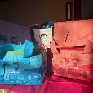 May include: Several Trader Joe's insulated bags in various colors are displayed. A blue mini insulated bag is in the foreground, with a green zipper and blue handles. Other bags are pink and purple, all featuring the Trader Joe's logo and palm tree graphics.