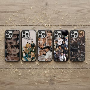 May include: Five black phone cases with different designs. The cases feature images of a musical group, with the group's name, "Stray Kids," printed on some cases. The cases are displayed on a wooden surface.