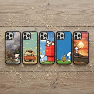 Può includere: Cinque custodie per telefoni con grafica a cartoni animati. Ogni custodia presenta una scena diversa con Snoopy e altri personaggi. Le custodie hanno bordi neri e sono esposte su una superficie di legno.