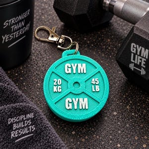 May include: A circular teal keychain shaped like a weight plate with a silver-toned lobster clasp. The plate features white embossed text reading GYM, 20 KG, and 45 LB. The design includes a central hole and raised radial spokes on the surface.