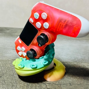 May include: A red and white video game controller resting on a teal, yellow, and white octopus-shaped stand. The controller has black joysticks and buttons with colored symbols. The stand has a gradient color scheme.