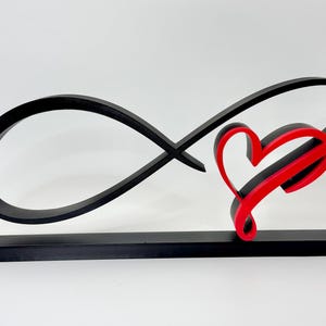 May include: A black infinity symbol sculpture with a red heart accent. The sculpture is mounted on a black rectangular base. The heart is outlined in black with a red interior. The design is modern and minimalist.