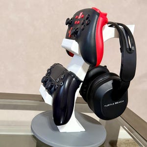 May include: A white and grey gaming accessory stand holding a black and red game controller, a black game controller, and black headphones. The headphones have the text "TURTLE BEACH" printed on them. The stand has a circular base.