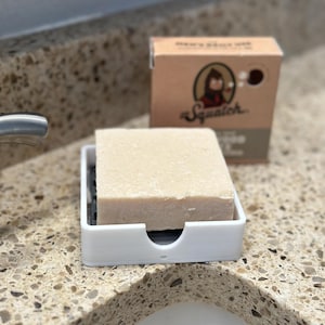 May include: A bar of soap rests in a white soap dish on a speckled countertop. The soap is light brown and rectangular. A cardboard box with the word "Squatch" and a cartoon image is in the background.