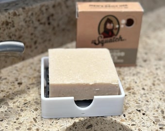 May include: A bar of soap rests in a white soap dish on a speckled countertop. The soap is light brown and rectangular. A cardboard box with the word "Squatch" and a cartoon image is in the background.