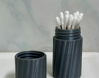 May include: A dark gray, cylindrical container with a matching lid. The container is filled with white cotton swabs. The container has a ridged, spiral design. The lid is next to the container.