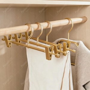 Pode incluir: Um conjunto de cabides para calças em metal dourado com clipes ajustáveis, suspensos numa barra de armário de madeira clara. Os cabides apresentam uma barra horizontal com dois clipes deslizantes para prender peças, com tecido branco.