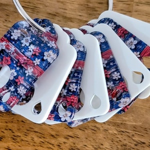 May include: A collection of white and patterned keychains on a silver ring. The patterned keychains feature a design of red, white, and blue with floral and star accents. The keychains have a teardrop-shaped hole.
