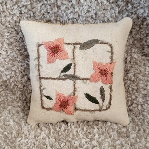 May include: A small, square decorative pillow with a cream-colored linen fabric. The pillow features an embroidered design of pink flowers, green leaves, and a brown square grid. The pillow is a home decor item.