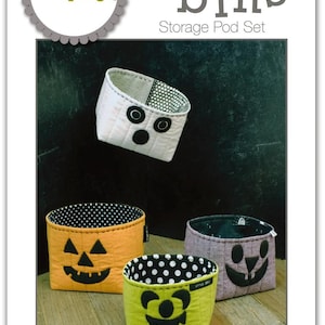 May include: A set of four fabric storage pods titled Boo Bins by Sue Spargo. The pods are white, orange, purple, and green, featuring stitched facial designs. Dimensions are listed as: Green 4.5x4.5x3.5 inches, Purple 5x5x4 inches, Orange 5.5x5.5x4.5 inches, White 5.5x5.5x3.5 inches.