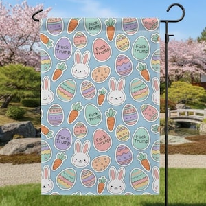May include: A light blue Easter garden flag featuring a repeating pattern of cartoon bunnies, carrots, and decorated eggs. The flag includes the text "Fuck Trump" on several of the egg-shaped designs.