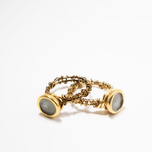 May include: Two gold-colored rings with a circular, light-colored stone setting. The bands have a textured, woven design. The rings are displayed on a white background, showcasing the jewelry's details.