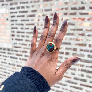 Puede incluir: Un anillo dorado con un centro redondo y estampado en tonos turquesa, rojo y negro. La banda del anillo presenta un diseño tejido. La mano que lleva el anillo tiene esmalte de uñas oscuro, sobre un fondo de pared de ladrillo.