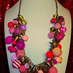 Könnte beinhalten: Eine farbenfrohe Statement-Halskette mit einer Vielzahl von rosa und roten Perlen. Die Kette hat eine Messingkette und dekorative Blattakzente. Die Perlen sind in verschiedenen Formen und Größen, darunter runde, gestreifte und strukturierte Designs.