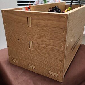 May include: A wooden box with a natural finish, featuring dovetail joints and screws. The box has a rectangular shape and is partially filled with various items. The box is sitting on a brown surface.