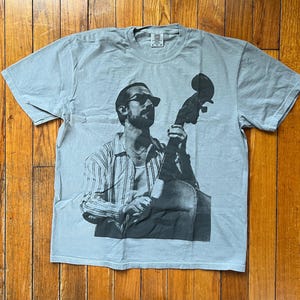 Könnte beinhalten: Hellblaues T-Shirt mit einem Schwarz-Weiß-Bild einer Person, die einen Kontrabass spielt. Die Person trägt eine Sonnenbrille und ein gestreiftes Hemd. Das T-Shirt ist aus weichem Material.