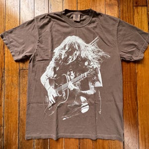 Puede incluir: Camiseta marrón con un gráfico blanco de un músico tocando una guitarra acústica. El gráfico muestra a una persona de pelo largo con la guitarra. La camiseta está hecha de un material suave.