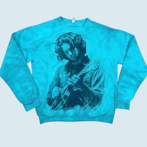 May include: A bright turquoise crewneck sweatshirt featuring a dark blue graphic print of a person with shoulder-length hair playing an acoustic guitar. The garment has long sleeves, a ribbed collar, and a mottled, tie-dye style fabric texture.
