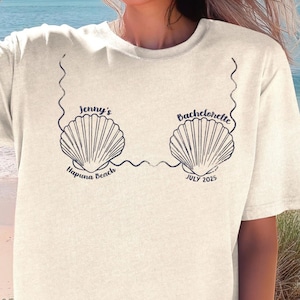 May include: Cream-colored t-shirt with a seashell design and the text "Jenny's Bachelorette Hapuna Beach JULY 2025". The shirt is worn with denim shorts on a beach setting.