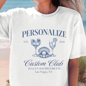 May include: White t-shirt featuring a blue graphic with the words "Personalize Custom Club", cocktail illustrations, and the text "Julia's Bachelorette, Las Vegas, NV". The shirt is worn on a beach.
