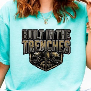 Könnte beinhalten: Hellblaues T-Shirt mit dem Text "BUILT IN THE TRENCHES" in einem schwarz-goldenen Design. Die Grafik zeigt zwei Football-Helme und eine Schaufel. Das Shirt ist aus einem weichen Material gefertigt.