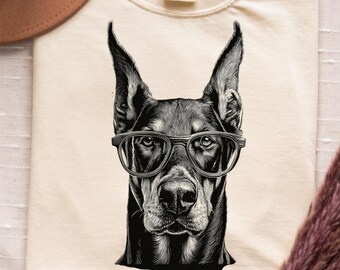 Può includere: T-shirt color crema con un'illustrazione in bianco e nero di un Dobermann con gli occhiali. Il testo "Doberman Dad" è stampato sotto l'immagine del cane. Un cappello marrone è visibile nell'angolo in alto a sinistra.