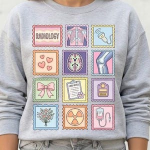 May include: A light gray sweatshirt with a colorful, postage stamp-style design. The stamps feature medical-themed illustrations and the word "RADIOLOGY". The design includes hearts, a brain scan, and a bow.