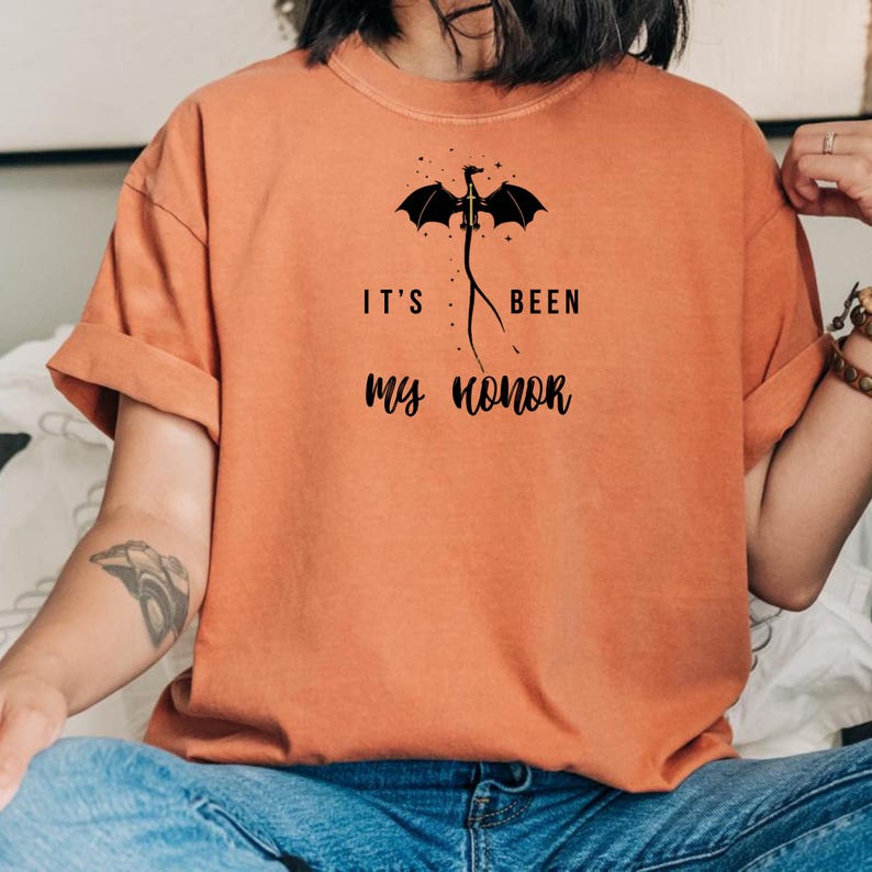 Puede incluir: Camiseta naranja con el texto "IT'S BEEN MY HONOR" y un gr&aacute;fico de un drag&oacute;n negro. El drag&oacute;n tiene las alas extendidas y est&aacute; posicionado encima del texto. La camiseta tiene mangas enrolladas y cuello redondo.