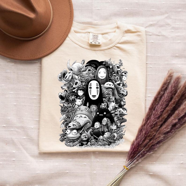 May include: Cream-colored t-shirt featuring a black and white graphic of various animated characters. The design includes characters from popular animated films. A brown hat and a bundle of dried flowers are also in the image.