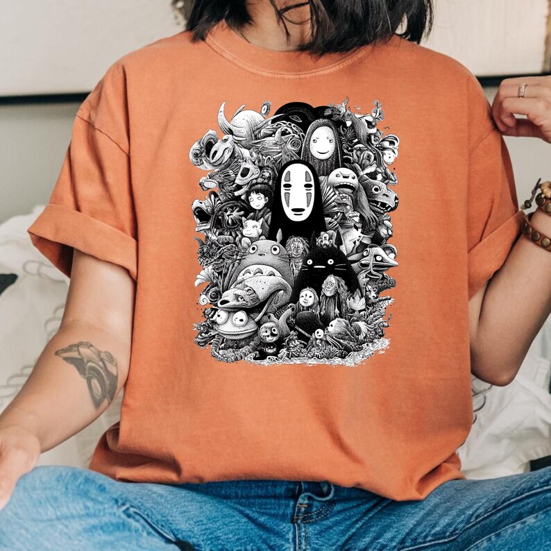 May include: An orange t-shirt featuring a black and white illustration of various animated characters. The graphic includes a central figure with a white mask and other creatures from the same animated universe.