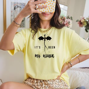 Puede incluir: Una camiseta amarilla clara con el texto "IT'S BEEN MY HONOR" y un gr&aacute;fico de drag&oacute;n negro. La camiseta es de estilo casual, adecuada para el uso diario. El dise&ntilde;o est&aacute; centrado en la parte delantera.