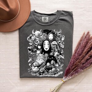 May include: Dark gray t-shirt featuring a black and white graphic of various animated characters. The design includes a central figure with a white mask and other whimsical creatures. The shirt is made of a soft material.