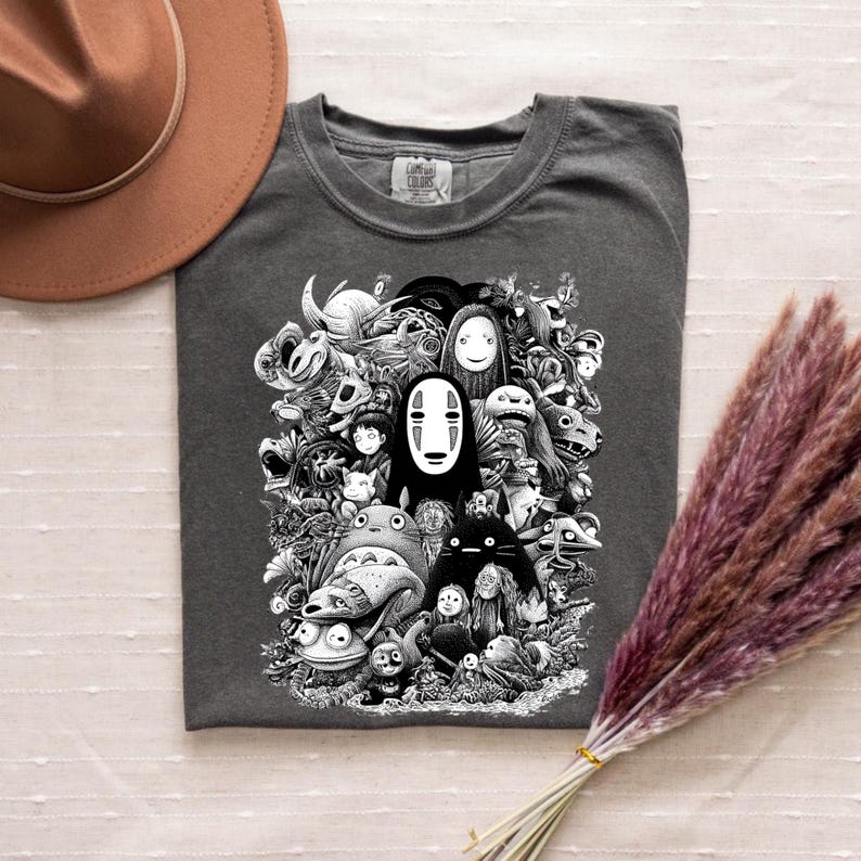 May include: Dark gray t-shirt featuring a black and white graphic of various animated characters. The design includes a central figure with a white mask and other whimsical creatures. The shirt is made of a soft material.