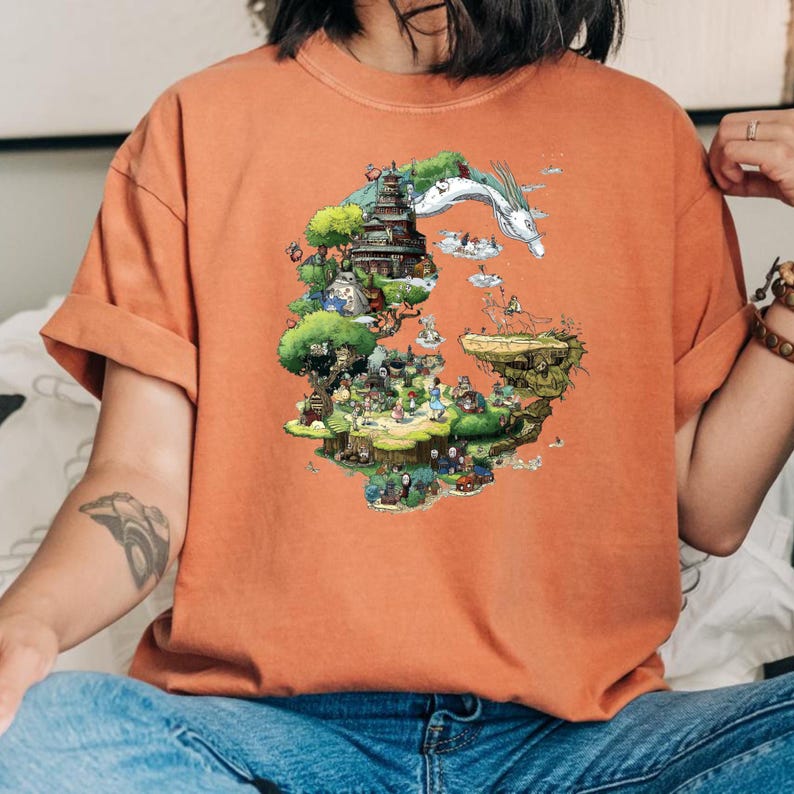 May include: An orange t-shirt featuring a colorful illustration of a fantasy landscape with a pagoda, a dragon, and various characters. The artwork is detailed and whimsical, with a variety of trees and buildings. The shirt has short sleeves and a crew neck.