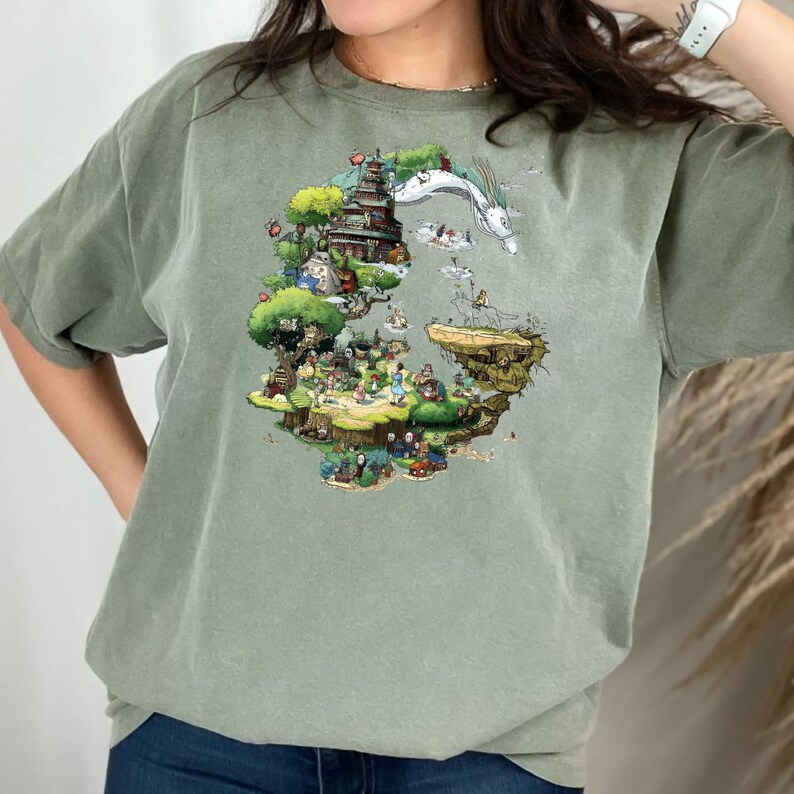 May include: Sage green t-shirt featuring a colorful illustration of a fantasy landscape with a pagoda, characters, and a dragon. The graphic is a detailed, whimsical design. The shirt has short sleeves and a crew neck.